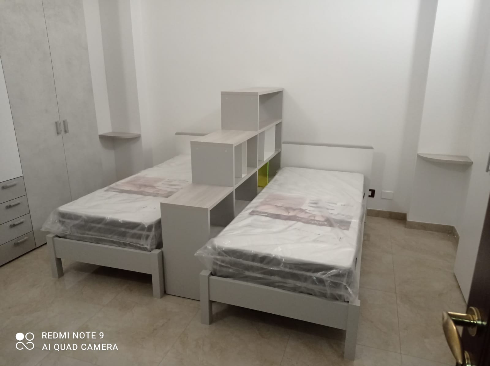 Due letti singoli con uno scaffale incorporato tra di loro, in una stanza prevalentemente bianca.