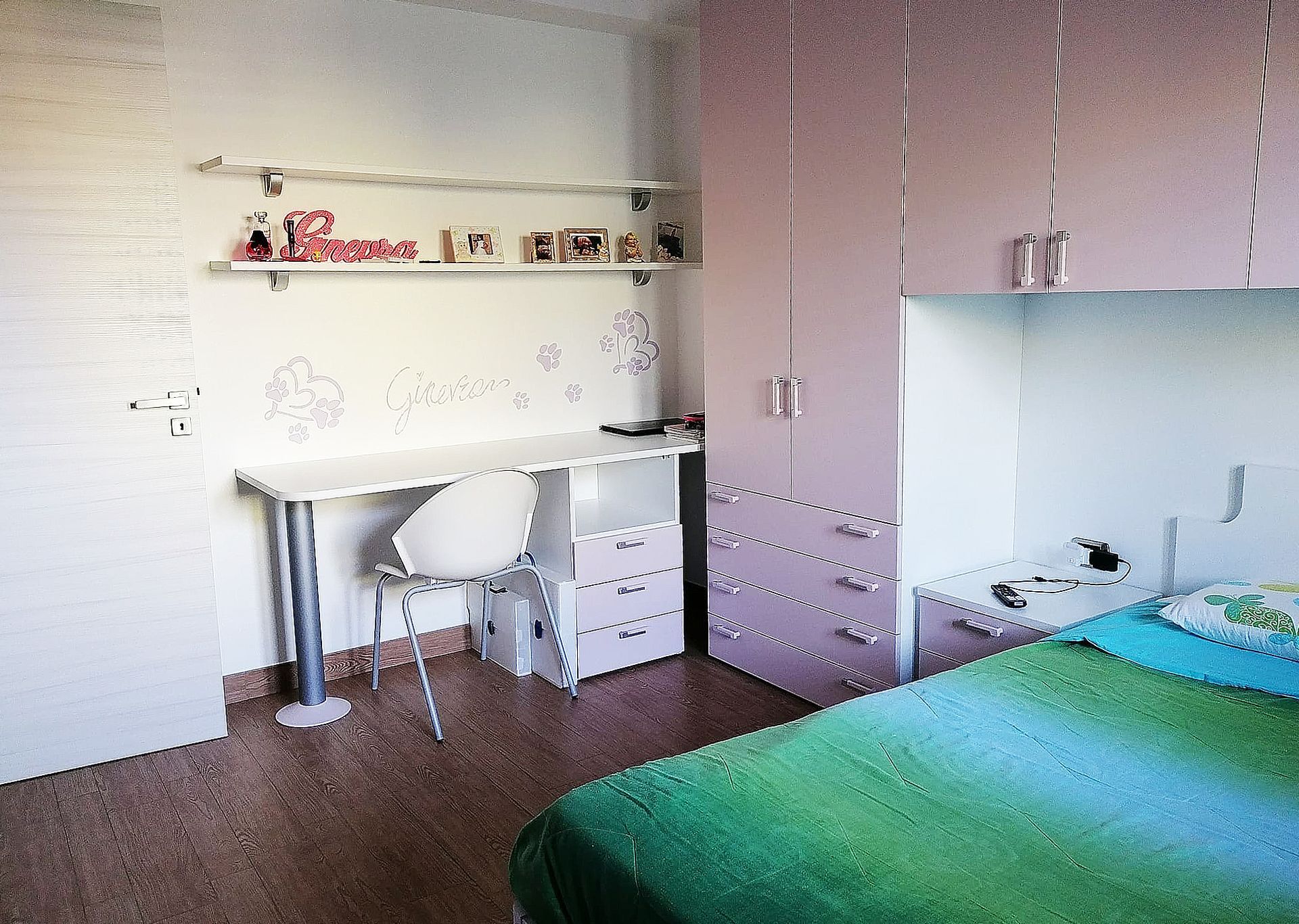 Camera da letto con scrivania, sedia, scaffali e un letto con lenzuola verdi e armadi rosa.