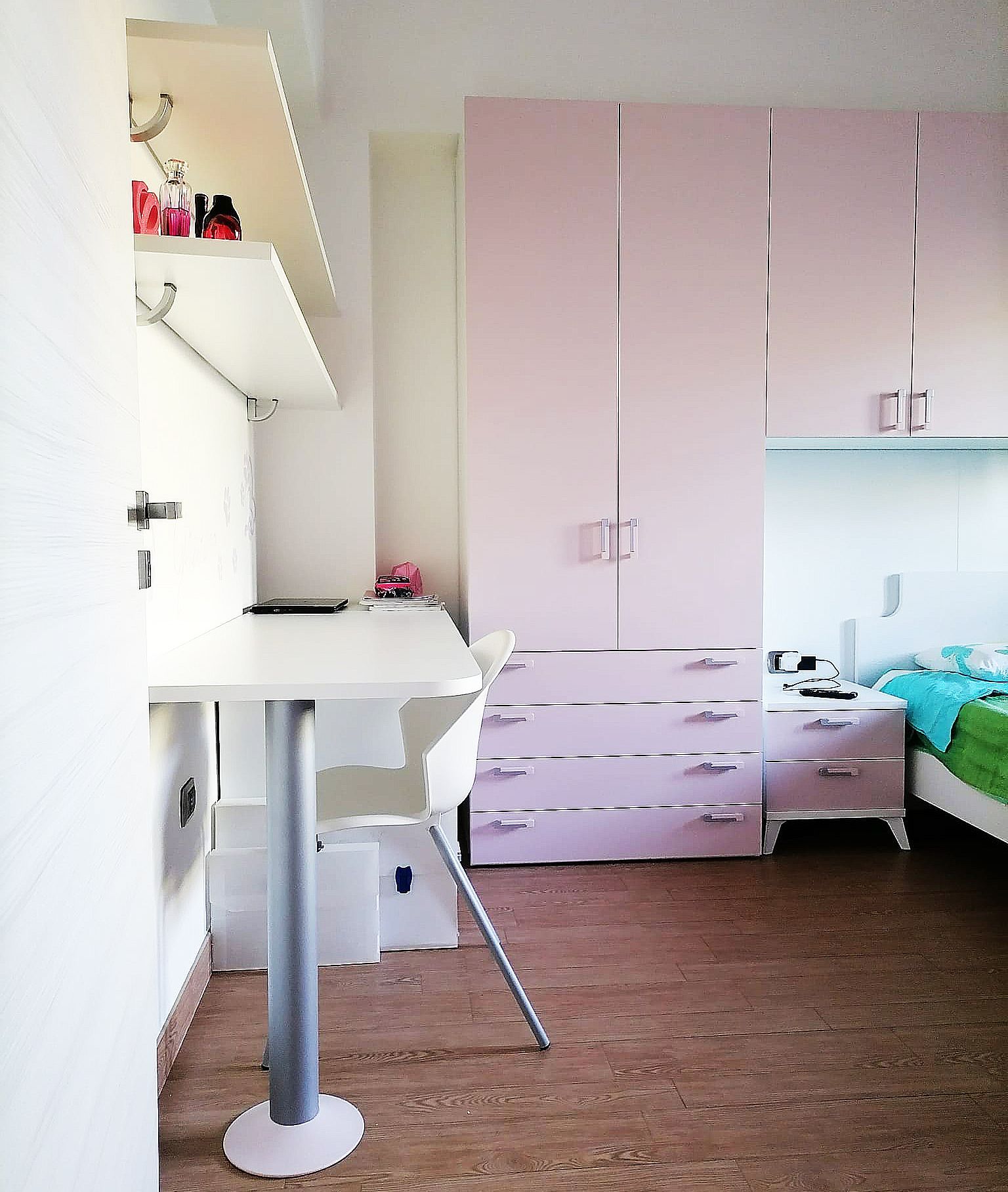 Una camera da letto rosa con scrivania, scaffali e armadio; una sedia bianca.