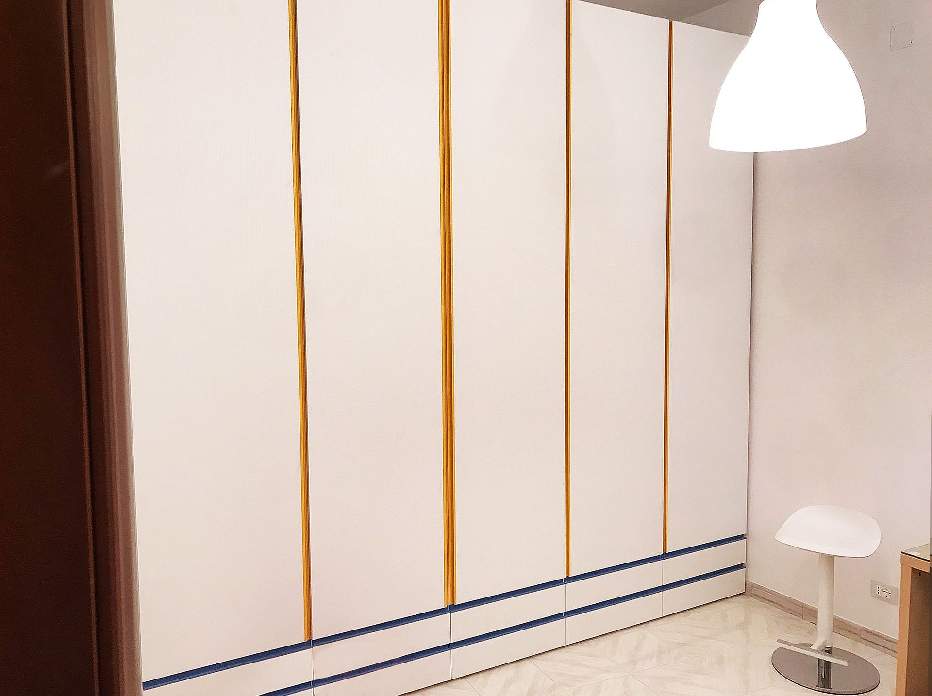 Armadio bianco con finiture dorate, accenti blu e lampada a sospensione in una stanza con uno sgabello.