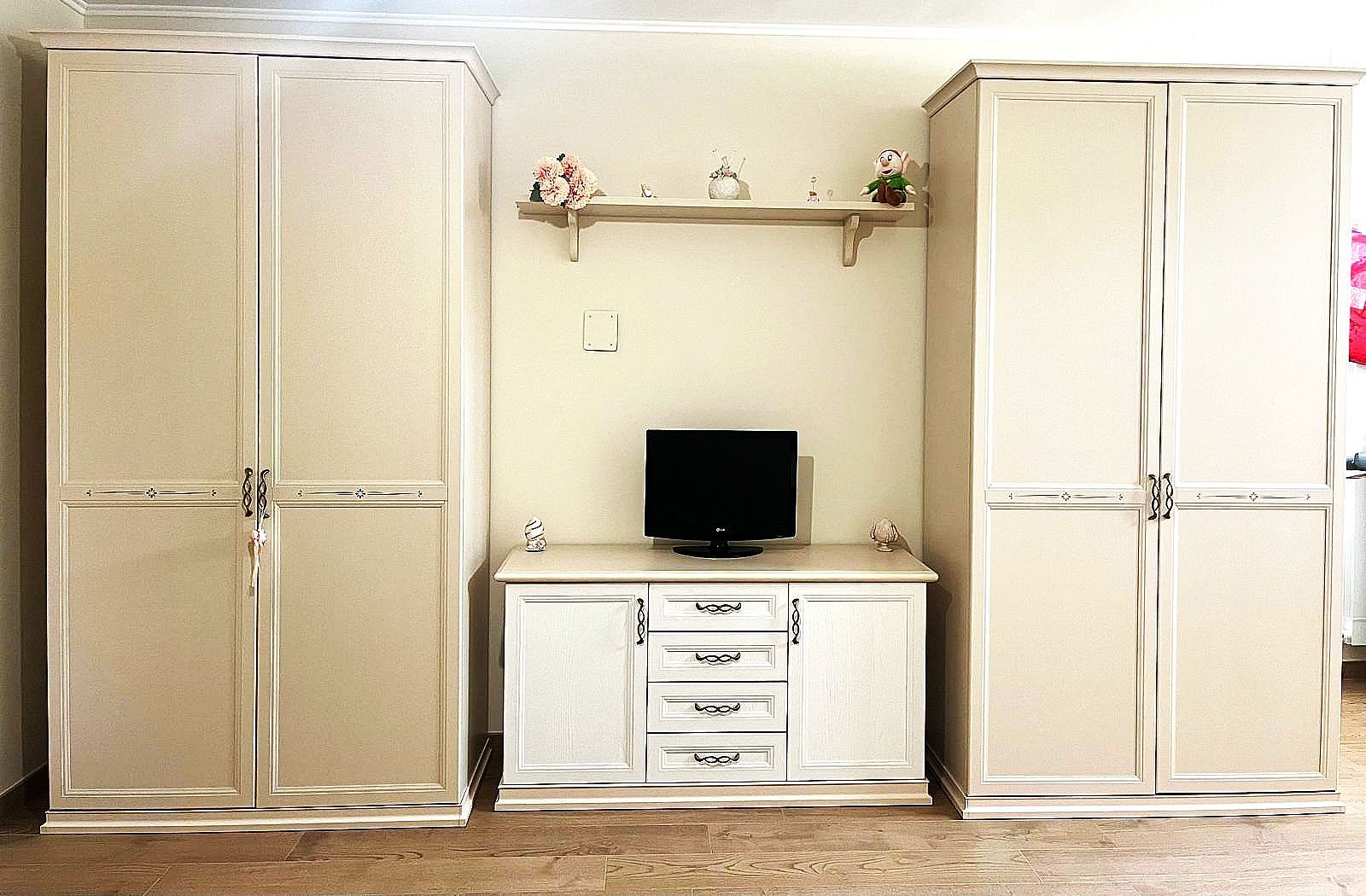 Set di mobili per la camera da letto: due alti armadi bianchi che fiancheggiano un mobile porta TV bianco con una piccola TV e uno scaffale con decorazioni.