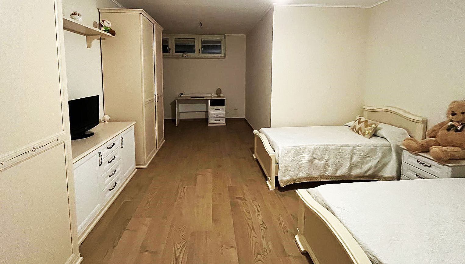 Camera da letto con due letti, ampia cassettiera con TV, scrivania e armadio. Colori beige e bianco.