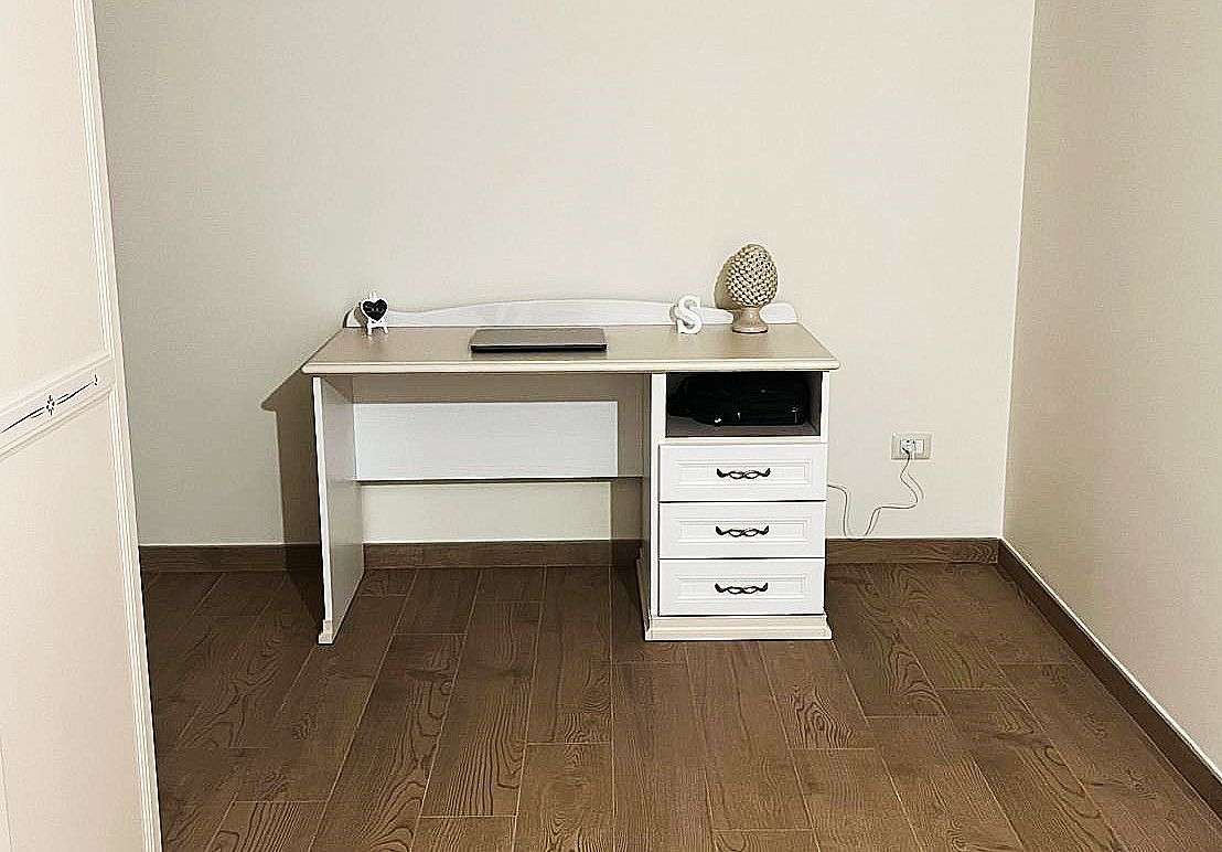 Scrivania in legno con cassetti contro una parete color crema, in una stanza con pavimenti in legno.