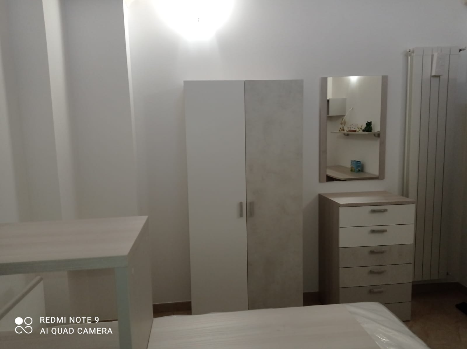 Camera da letto con mobili bianchi e beige: armadio, cassettiera, specchio e scrivania. Un termosifone si trova sulla destra.
