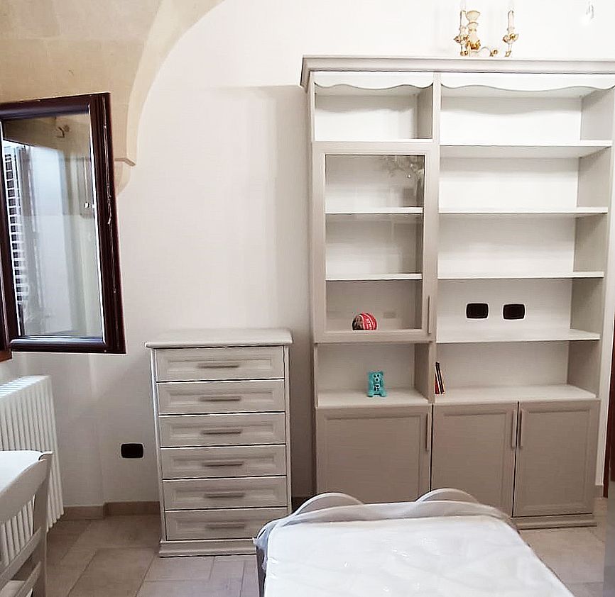 Camera da letto con cassettiera bianca, libreria, letto e finestra con finiture scure.