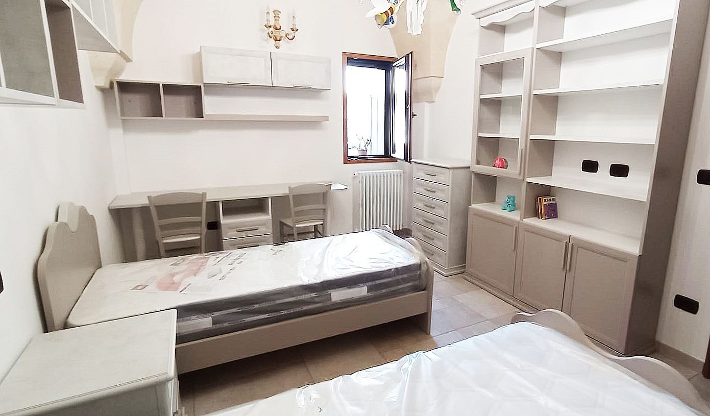 Camera da letto con due letti singoli, scrivanie, scaffali e una finestra. I colori chiari dominano la stanza.