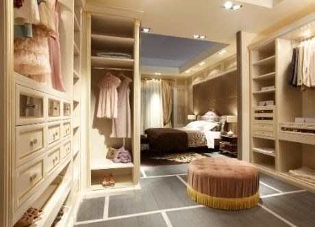 Cabina armadio con mobili beige, vestiti, cassetti, tappeto e una camera da letto visibile sullo sfondo.