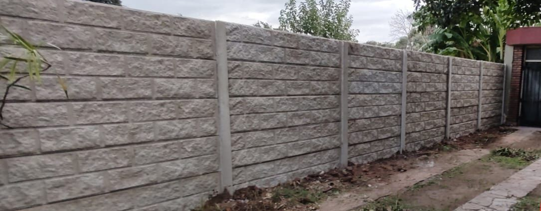Muro de bloques de hormigón junto a una acera y un camino de tierra, con árboles y una casa al fondo.