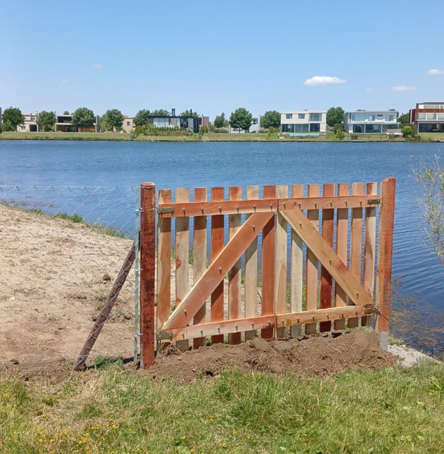 Una puerta de madera junto a un lago, con agua, césped y casas al otro lado de la orilla