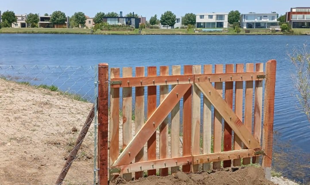 Puerta de madera junto a una orilla arenosa, con vistas a un lago tranquilo y casas al otro lado del agua
