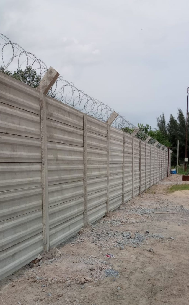 Un largo muro de hormigón coronado con alambre de púas junto a un camino de tierra, con una bolsa roja desechada en primer plano