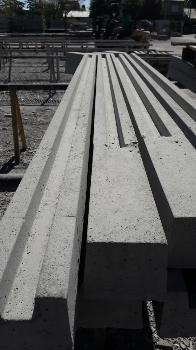 Vigas largas de hormigón apiladas en un patio de obras soleado.