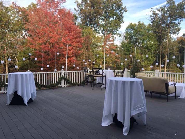 Outdoor deck set for an event with white tablecloths, hanging decorations, and fall foliage.
