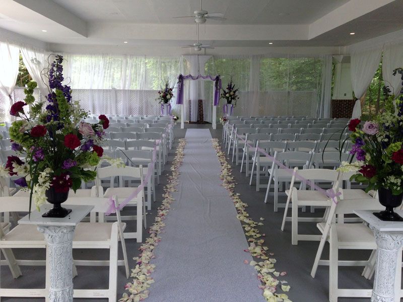 Wedding ceremony setup: white chairs, aisle with petals, floral arrangements, and archway with purple accents.