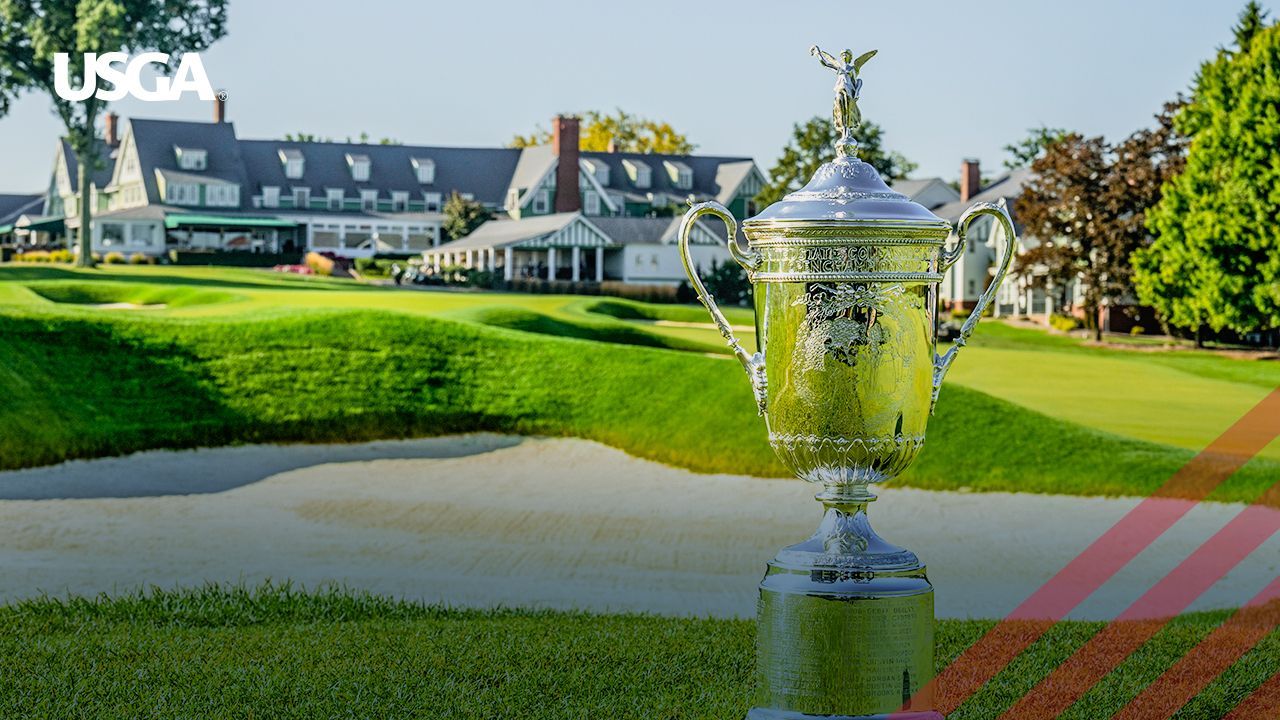 US Open trophy on a golf course, with a clubhouse in the background. Green grass and sand trap.