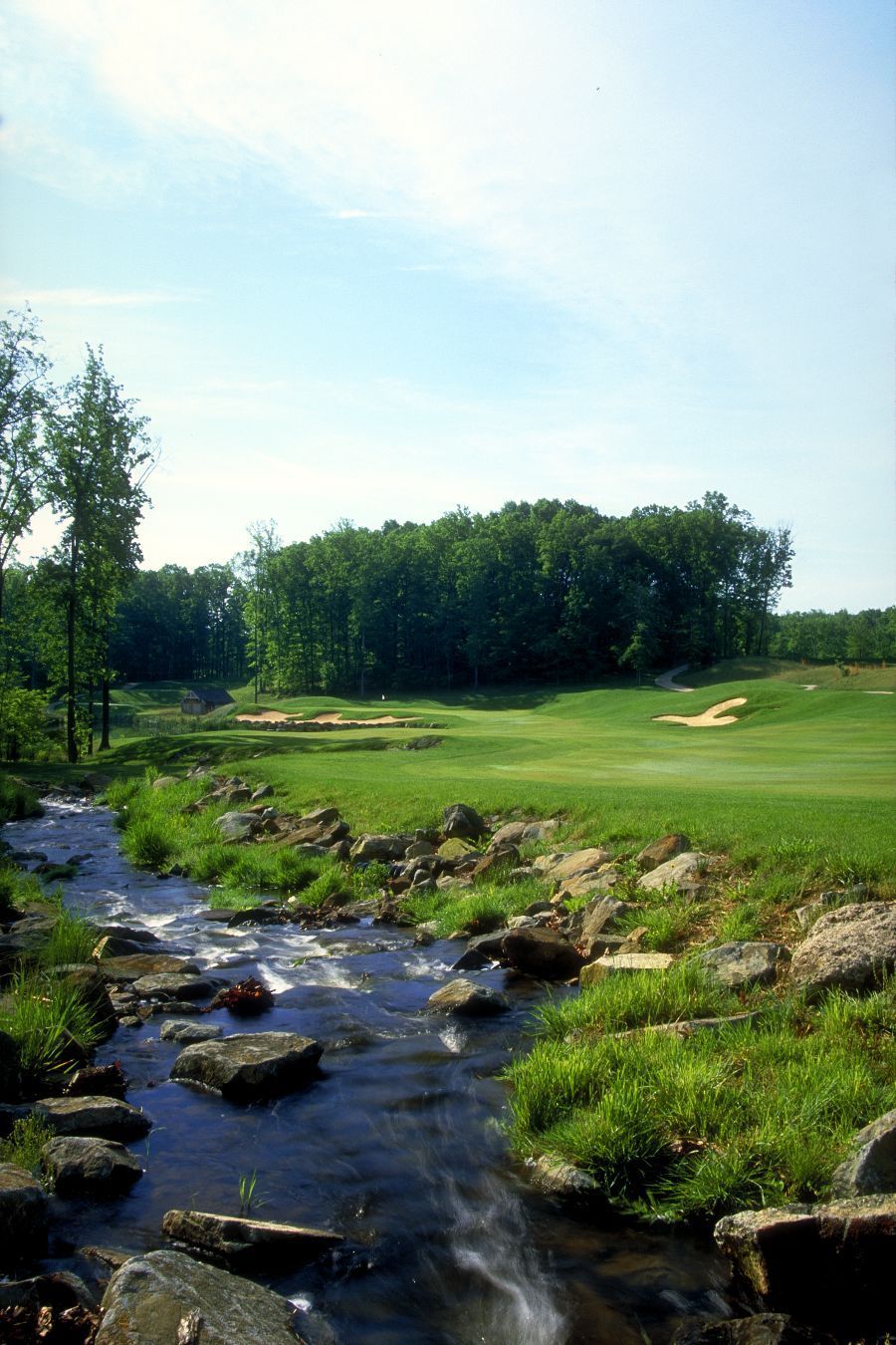 A golf course with a stream, green grass, trees, and a blue sky.