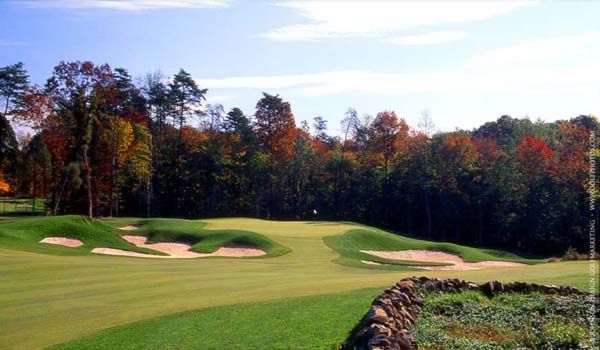 Golf course green with bunkers, trees with autumn colors, and a stone wall.