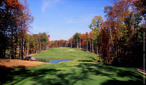 Golf course fairway surrounded by trees with autumn foliage, pond in the foreground, and blue sky.
