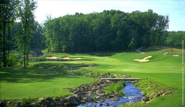Green golf course with a stream, bridge, and sand traps, surrounded by trees.