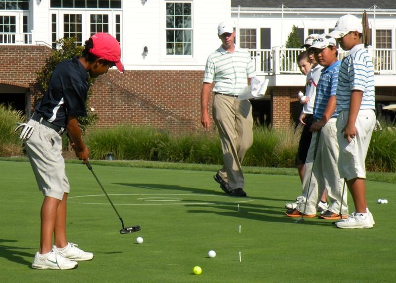 Boy in red cap putts on green; others watch, instructor in background.