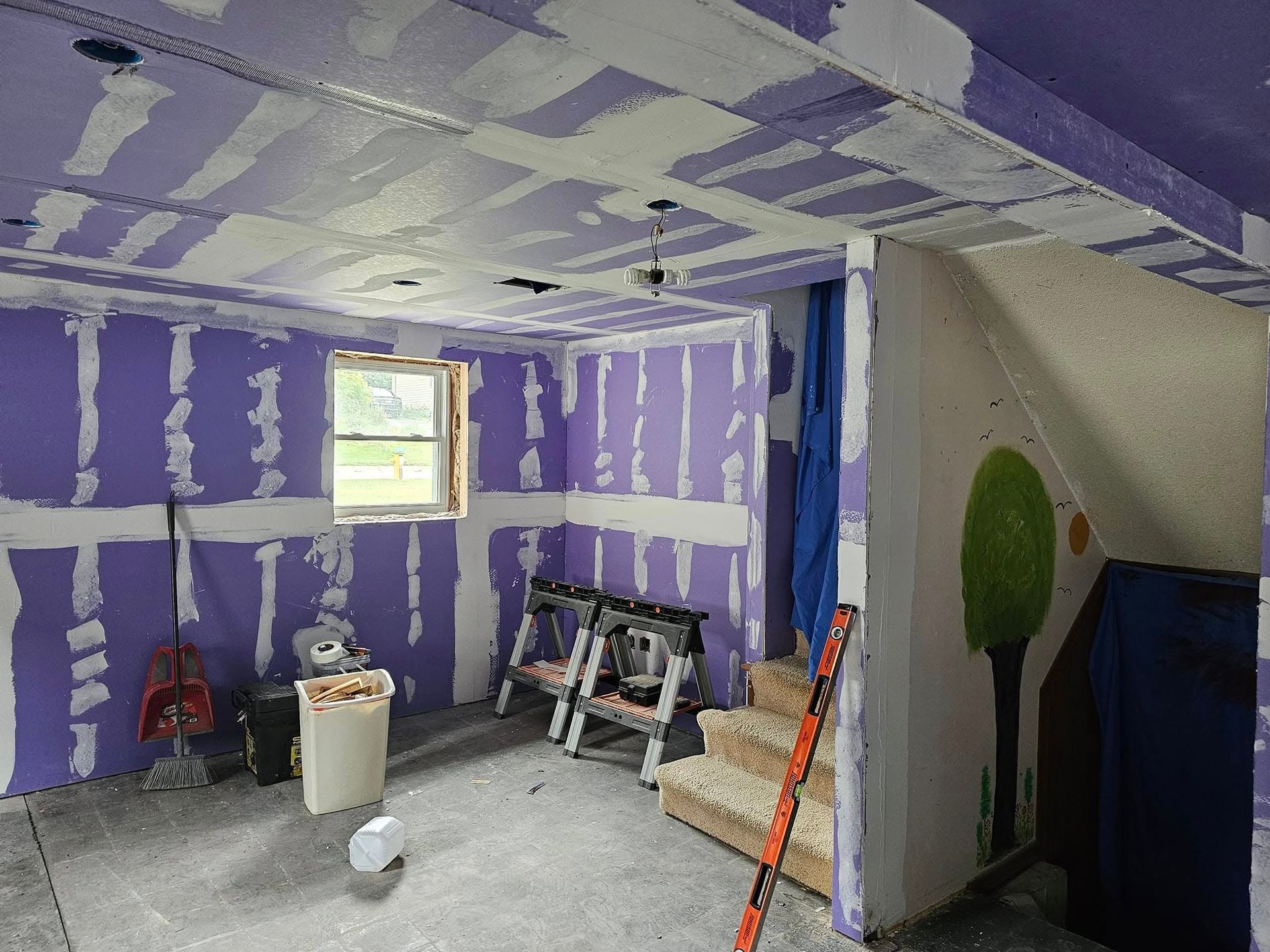 Interior room under construction, with purple drywall installed. Sawhorses, tools, and a window are visible.
