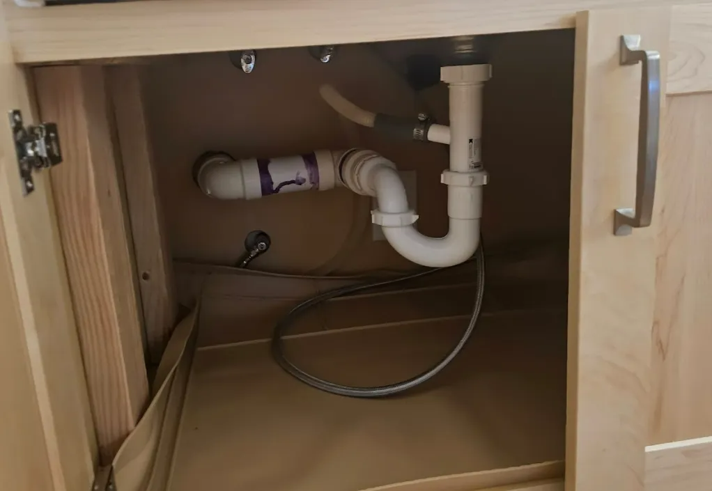 View inside a light-colored wooden cabinet under a sink, showing white plumbing and a gray corrugated drain pipe.