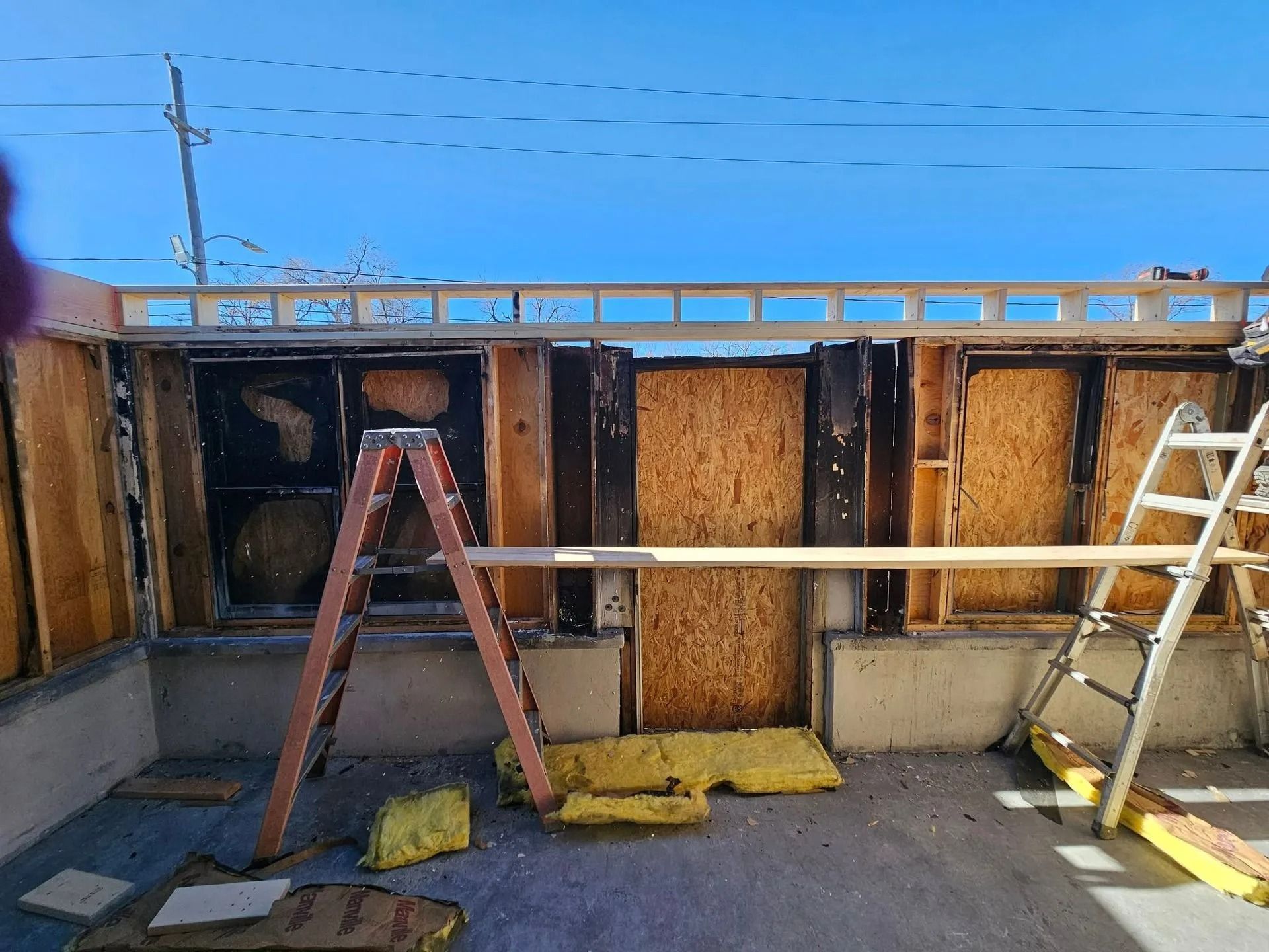 Construction site with exposed wall framing, plywood, insulation, and ladders against blue sky.