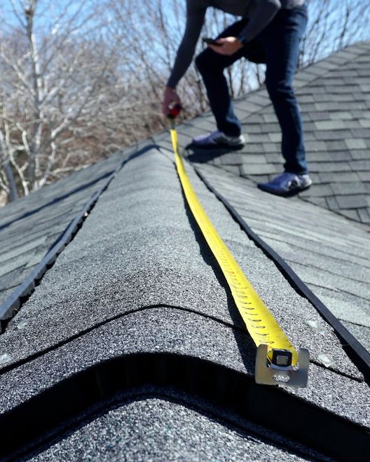 Person measuring a roof ridge with a yellow tape measure on gray shingles