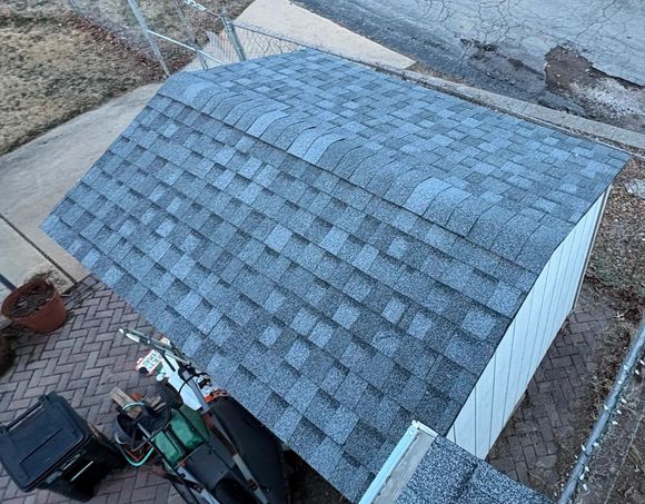 Overhead view of a gray shingled shed roof beside a driveway and brick patio, with lawn equipment nearby