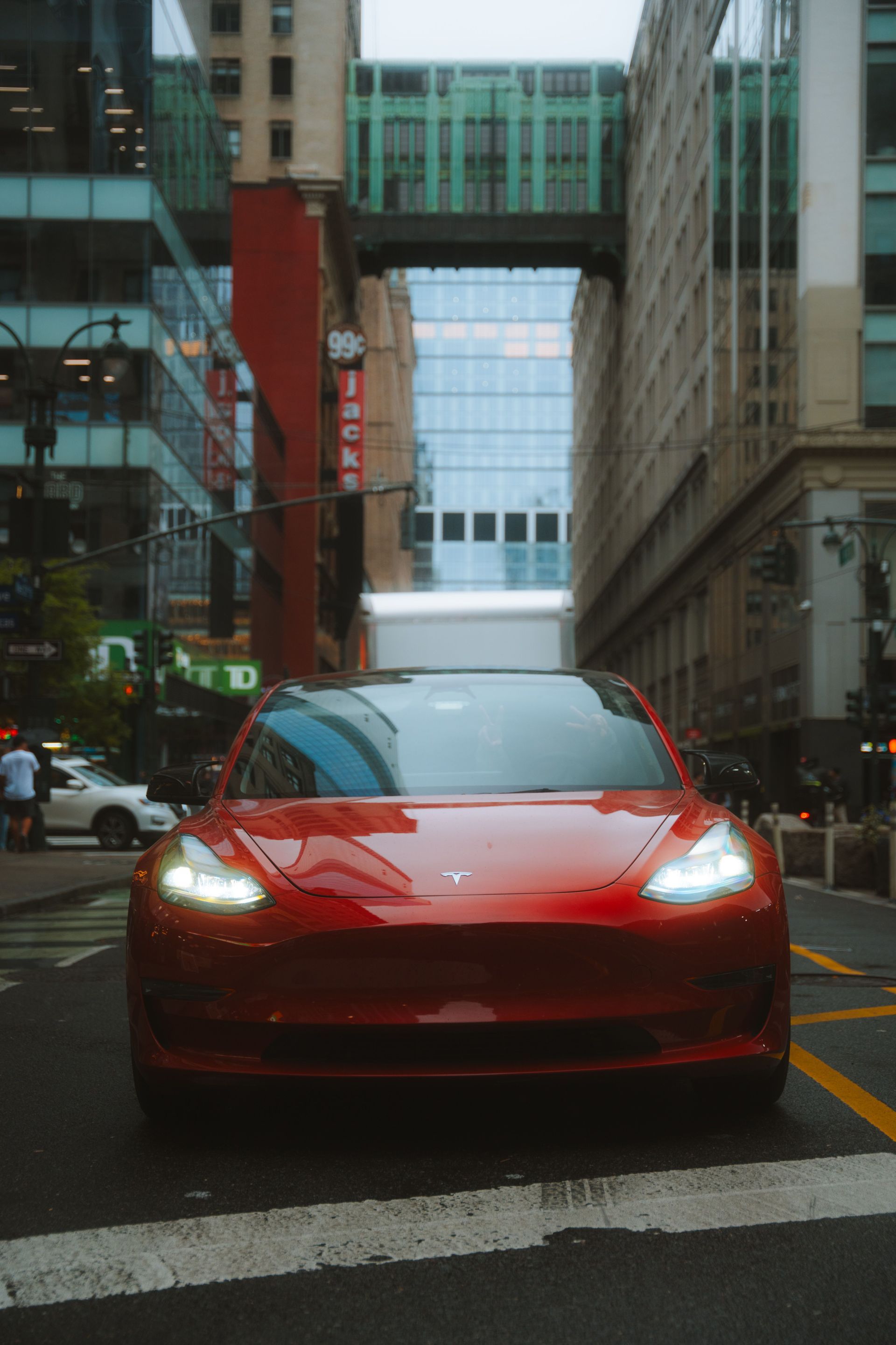 Red sports car driving on a city street between tall buildings, headlights on in the rain.