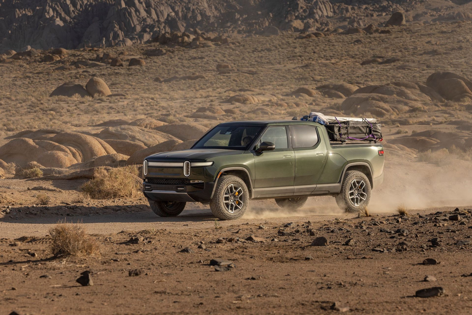 Green pickup truck driving through a dusty desert landscape with rocky hills in the background
