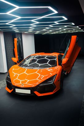 Orange sports car with raised scissor doors in a showroom with geometric ceiling lights