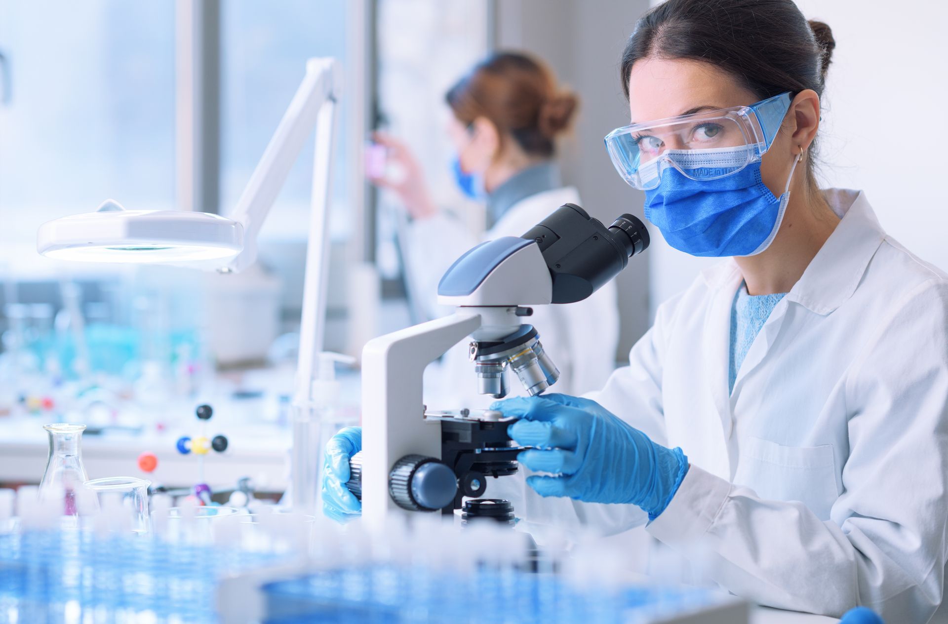 Científico con bata de laboratorio, máscara y gafas protectoras mirando a través de un microscopio; otra persona en el fondo.