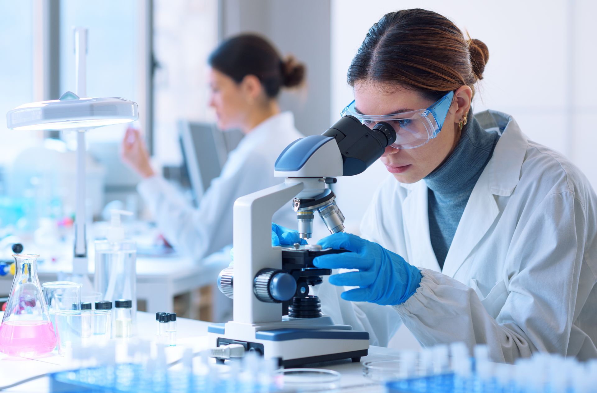 Una mujer con bata de laboratorio mira a través del microscopio, con guantes y gafas protectoras, y otro científico al fondo.