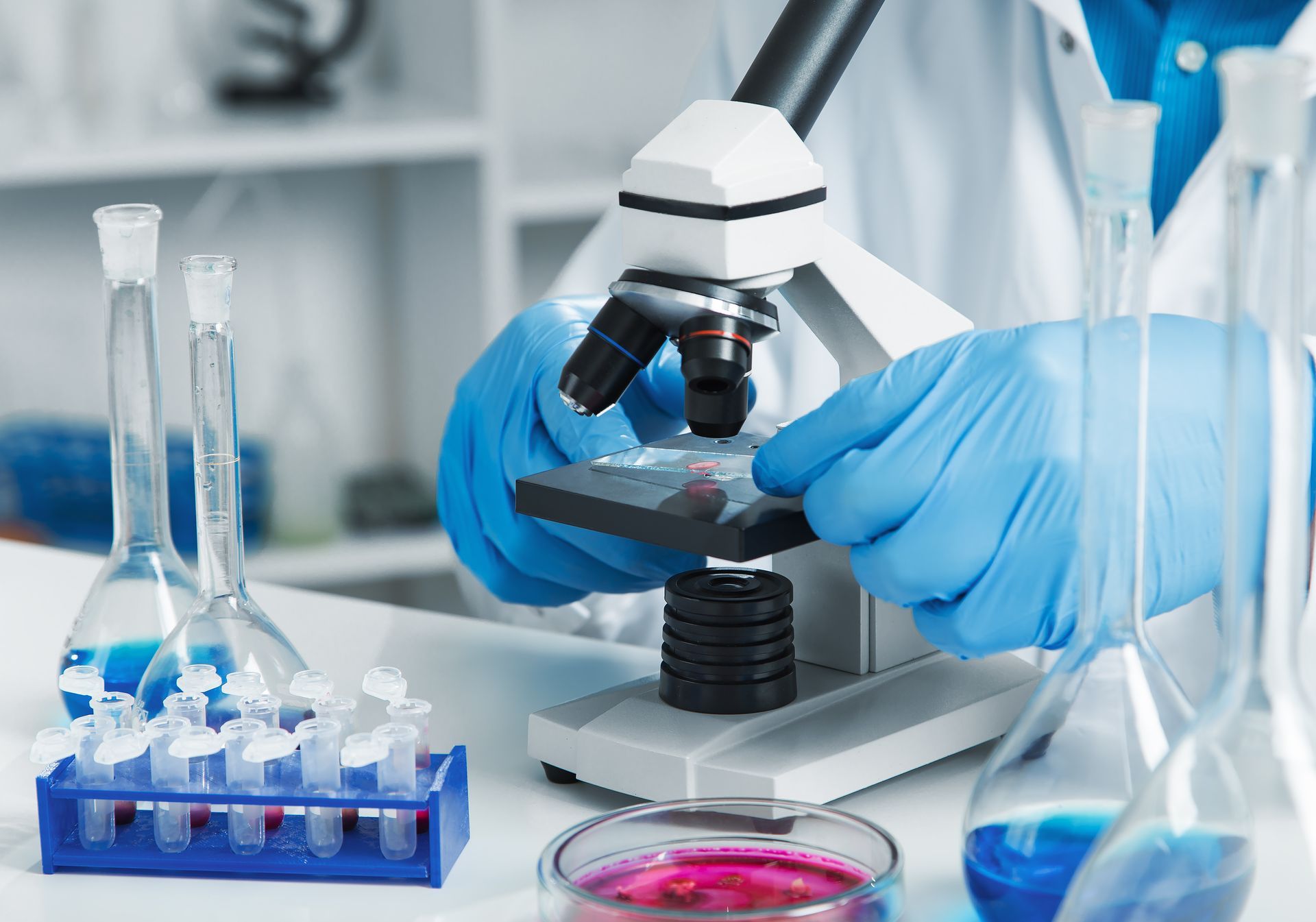 Persona con guantes azules utilizando un microscopio en un laboratorio, con matraces y tubos de ensayo visibles.