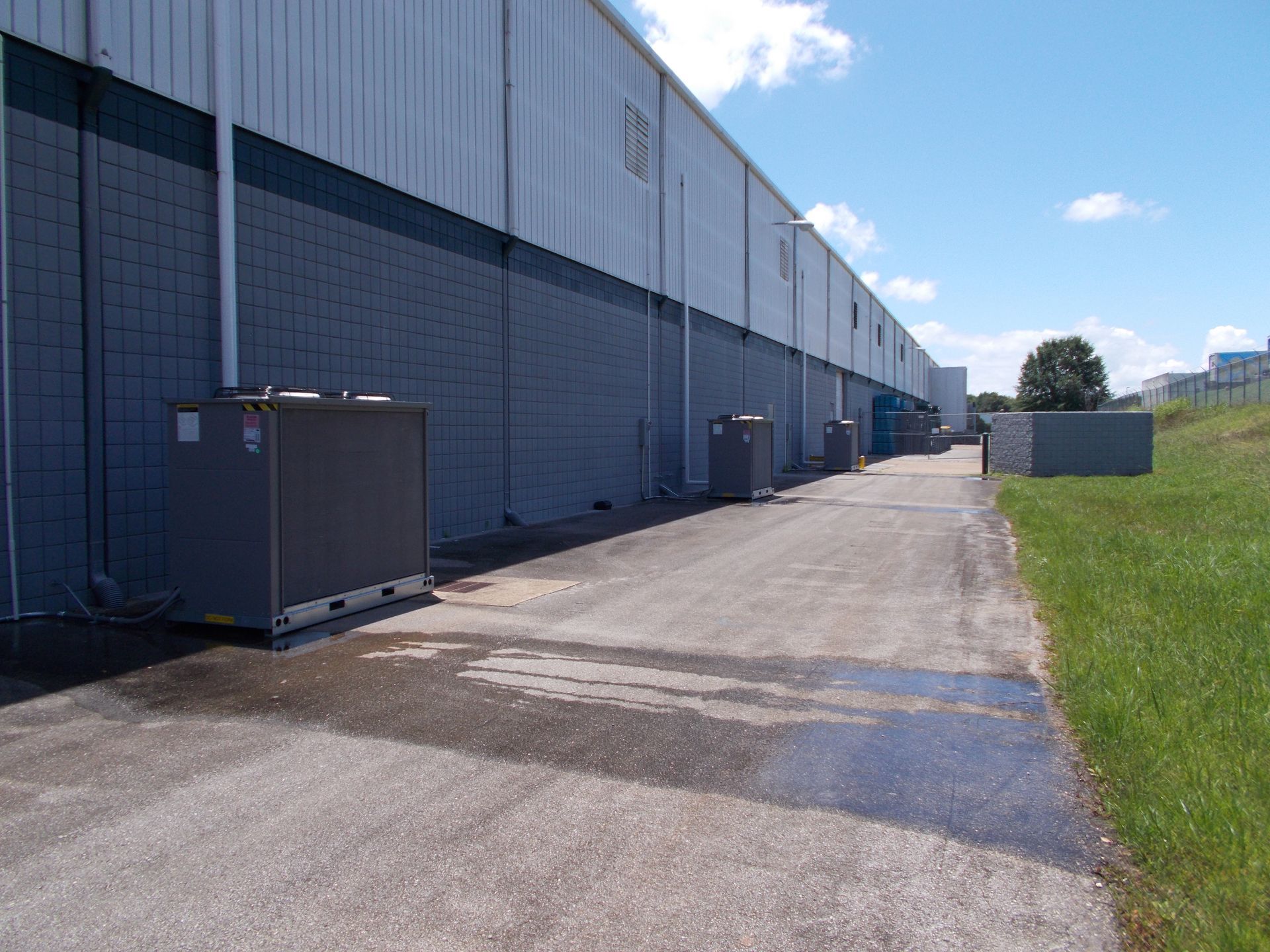Exterior side of a large gray warehouse with loading area, paved lane, and grassy strip under a blue sky