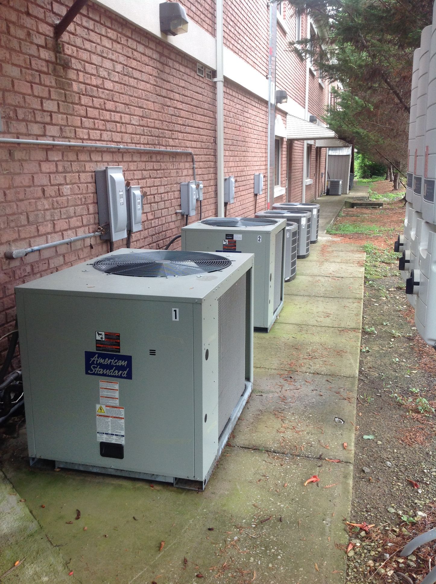 Row of outdoor HVAC units beside a brick building on a narrow walkway