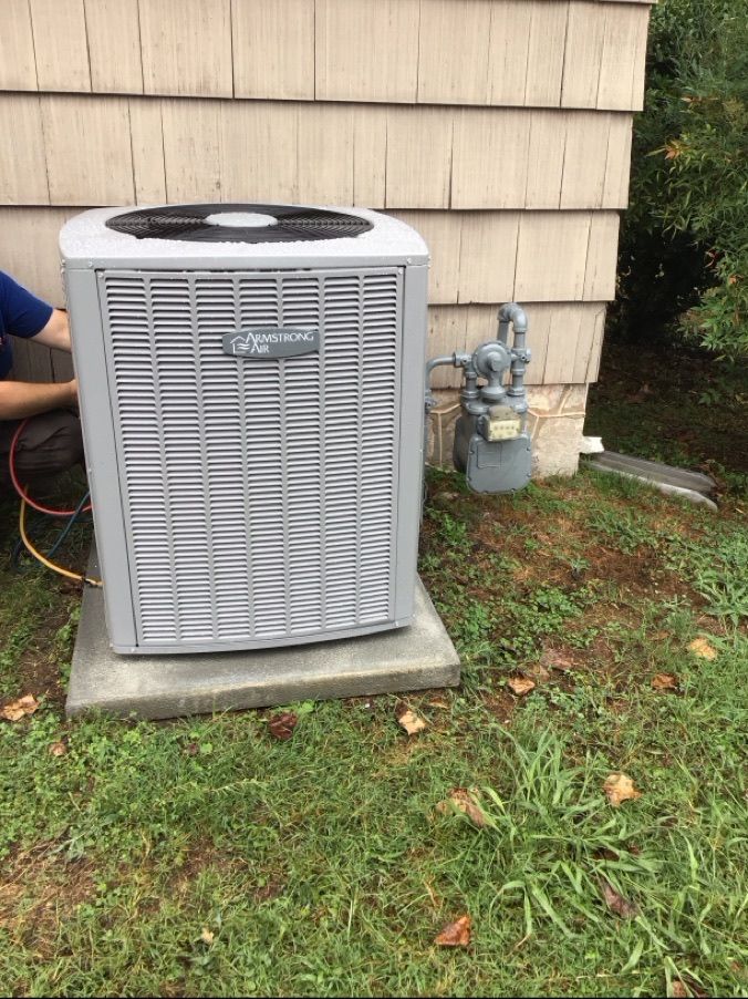 Gray outdoor air conditioner unit beside beige house siding on a concrete pad, with grass and utility pipes nearby