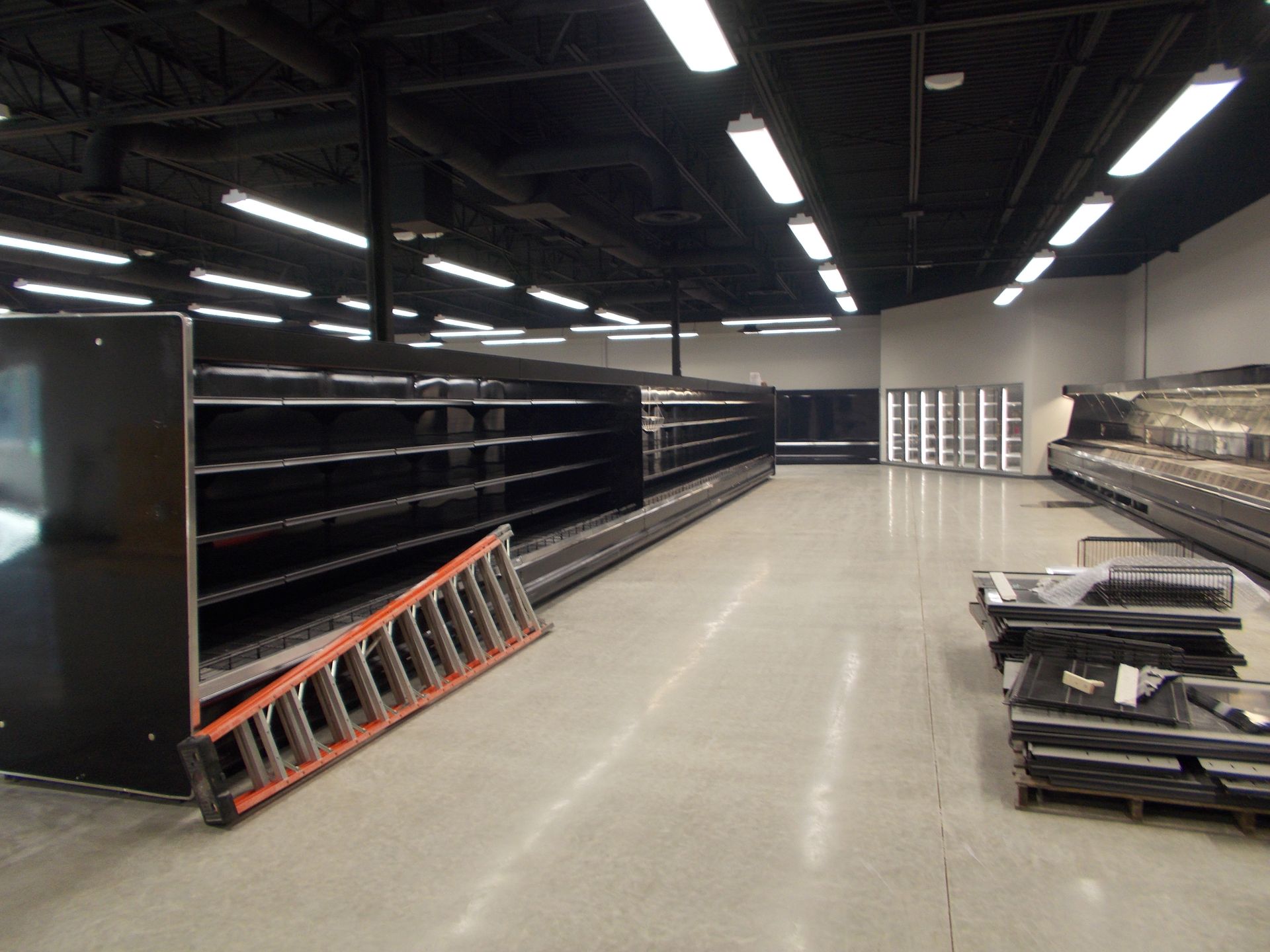 Empty warehouse aisle with long black shelving, orange ladder, and stacked pallets under bright ceiling lights