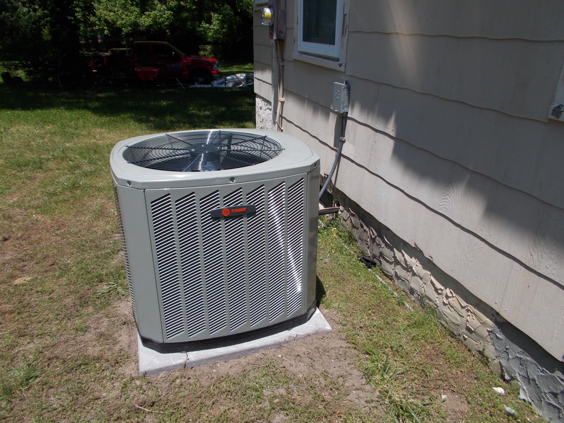 Outdoor HVAC condenser unit beside a house wall on a concrete pad, in a grassy yard.