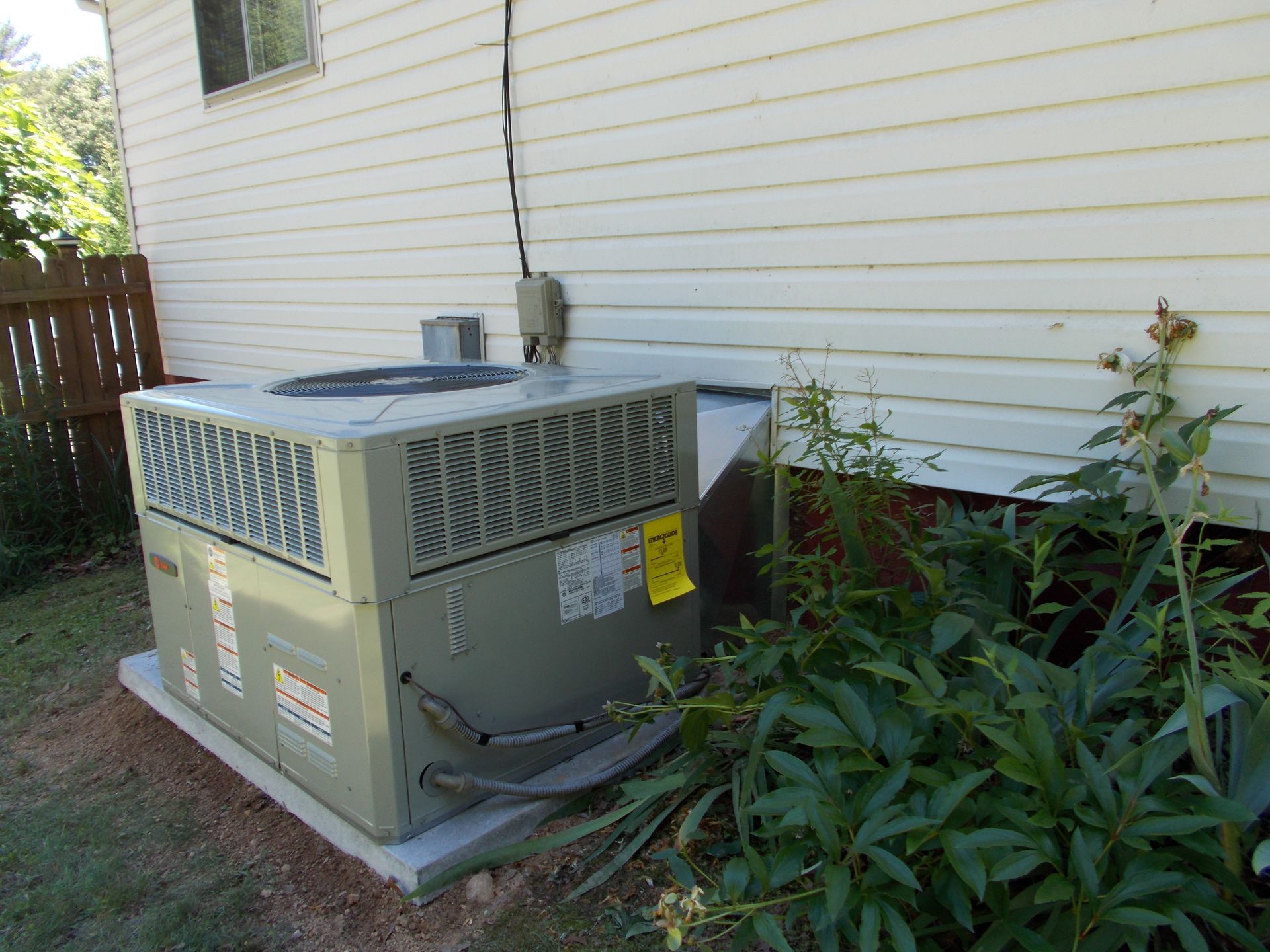 Outdoor air conditioner unit beside a white house wall and overgrown plants