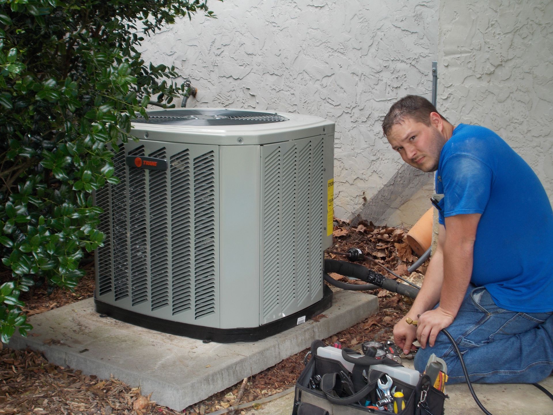 Technician kneeling beside outdoor air conditioner, using tools to repair the unit.