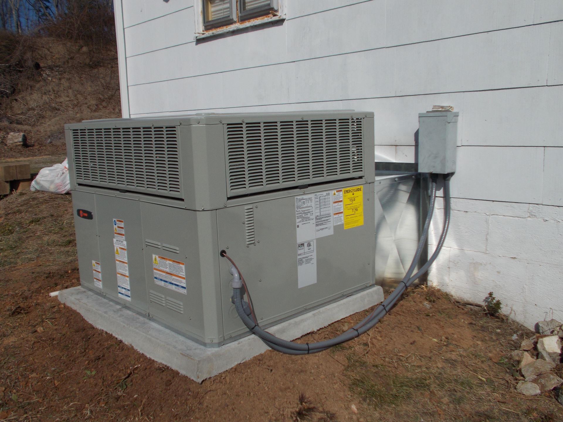 Outdoor HVAC unit beside a house, with visible electrical conduit and cables on a concrete pad.