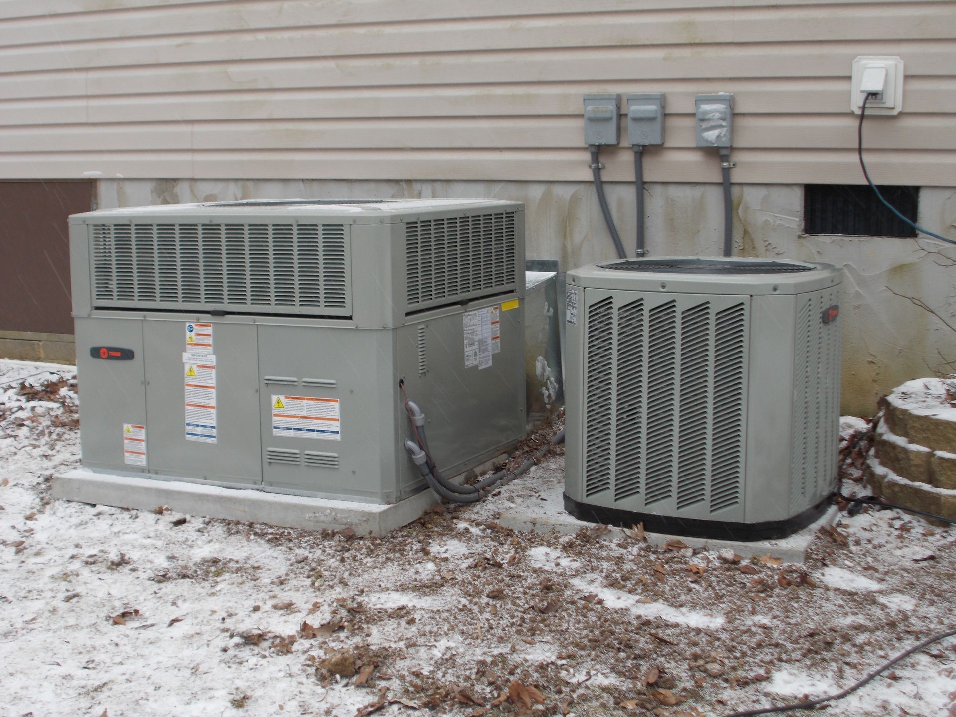 Two outdoor HVAC units beside a house wall on a snowy ground
