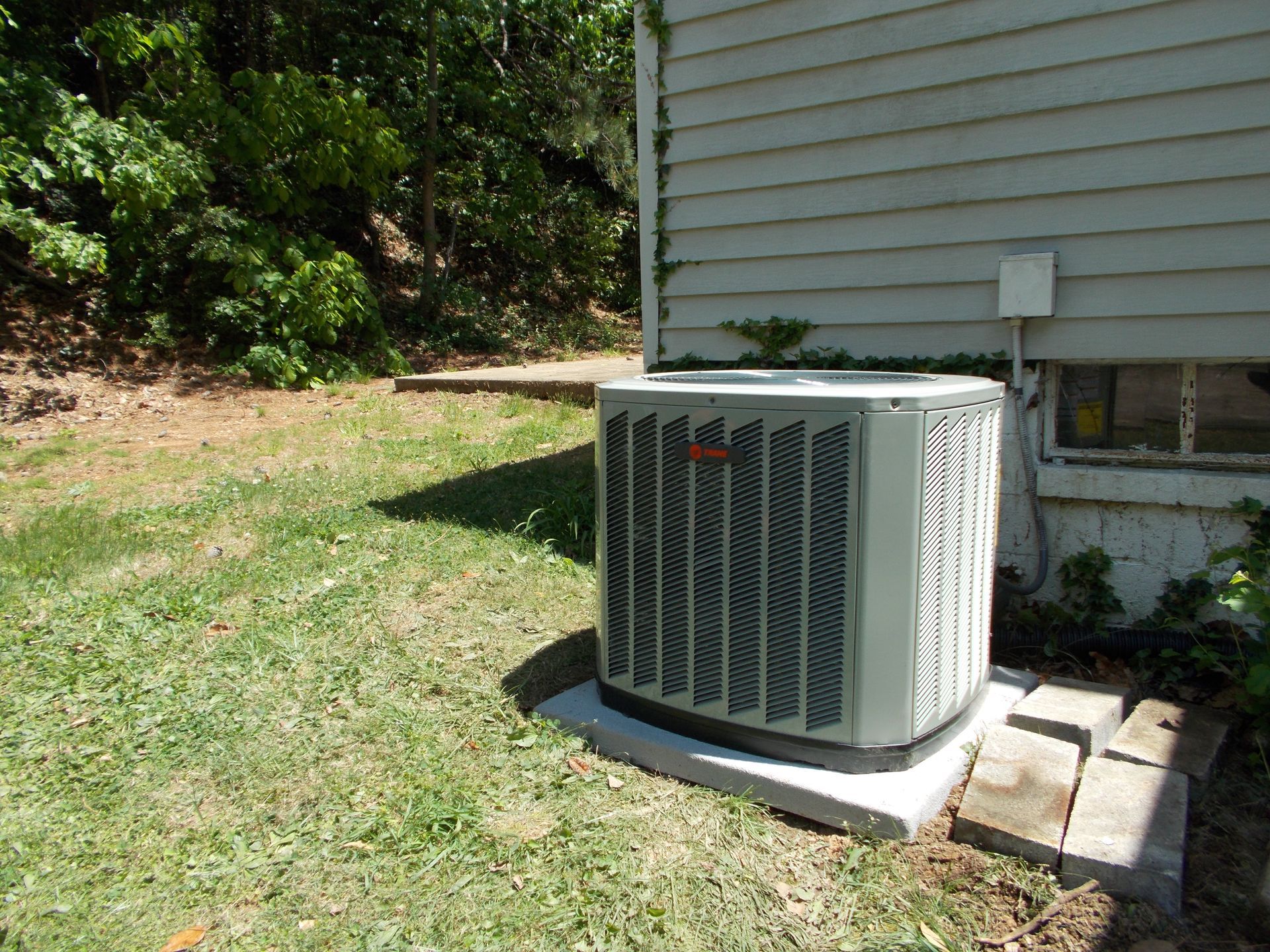 Outdoor air conditioner unit beside a house on a grassy lawn, near trees and a concrete pad