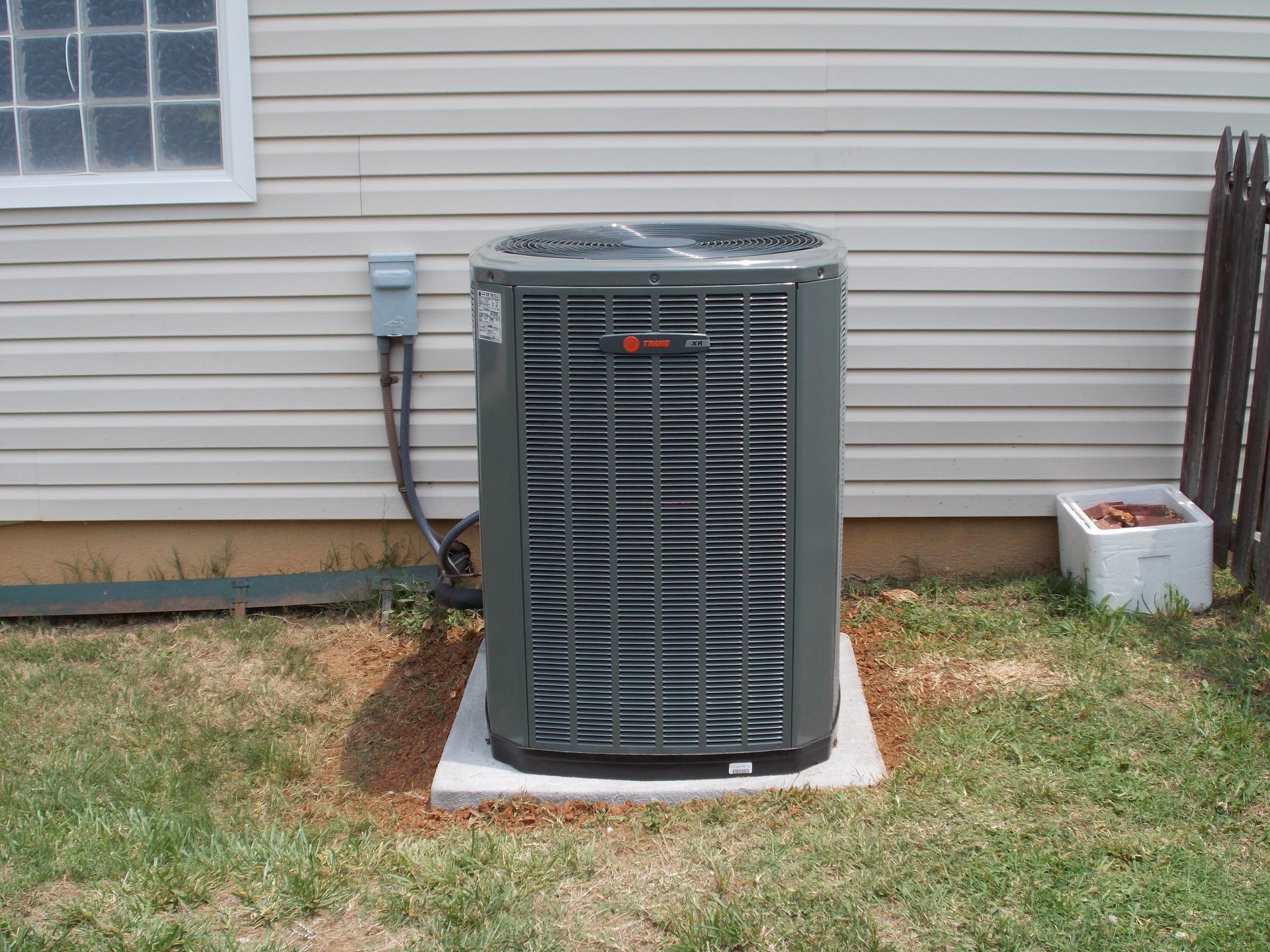Outdoor air conditioning unit beside a house wall, with grass and a small planter nearby.