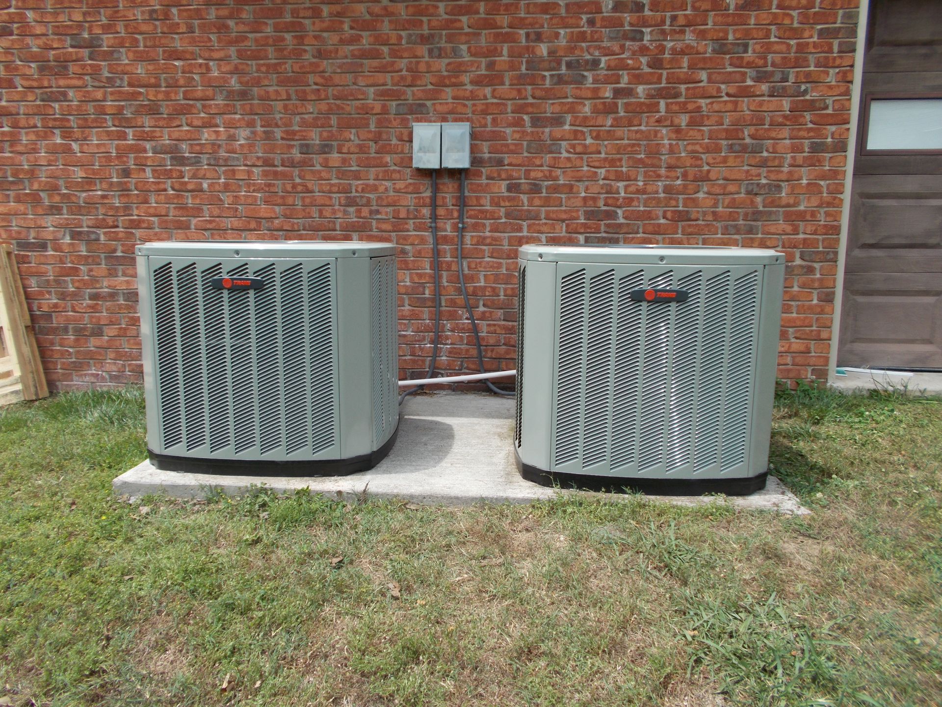 Two gray outdoor HVAC units beside a brick wall above a grass lawn
