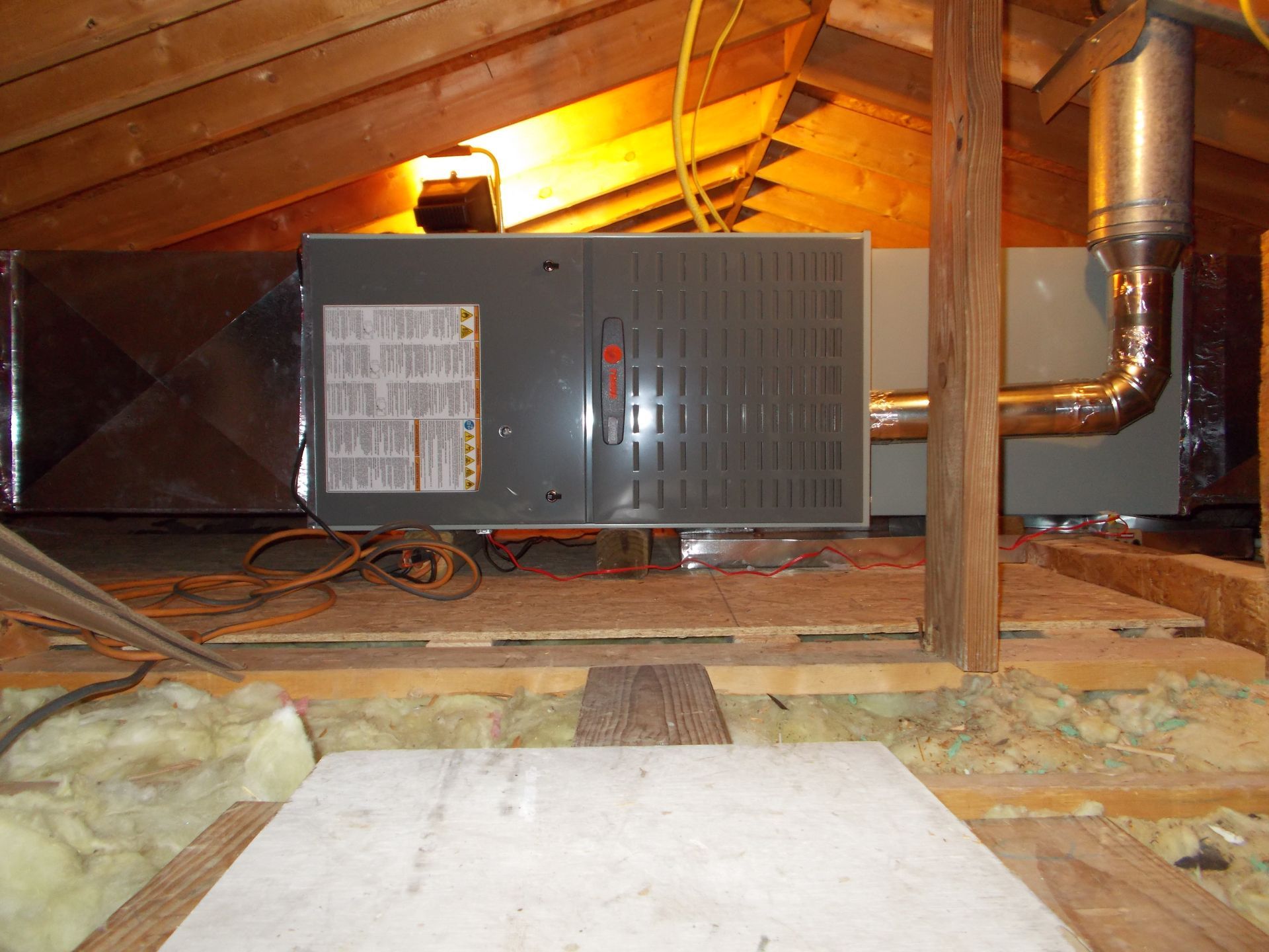 Attic with a gray furnace unit, exposed ductwork, and insulation under slanted wooden rafters.