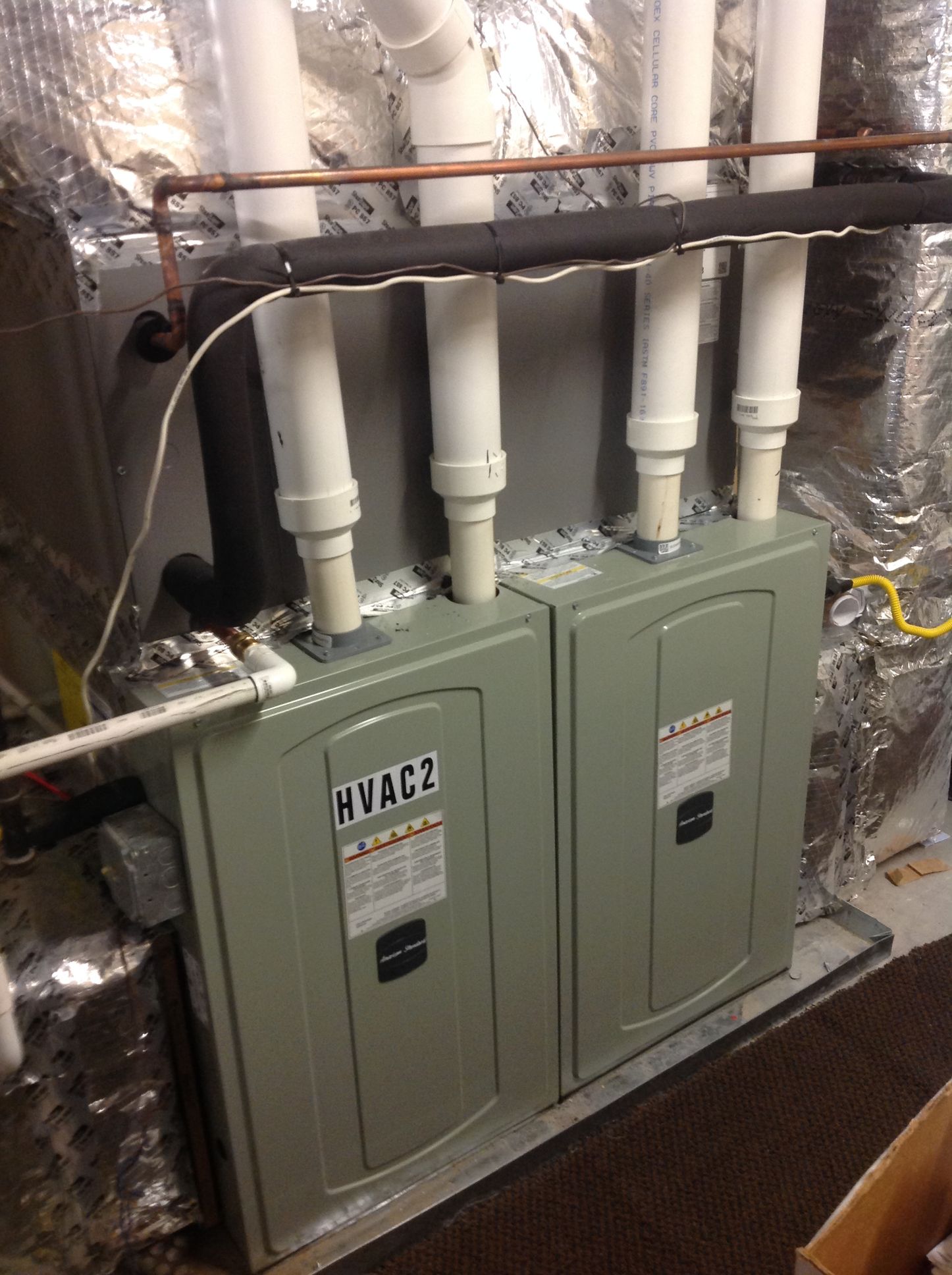 Two gray HVAC units with white pipes in a basement utility room covered in foil insulation
