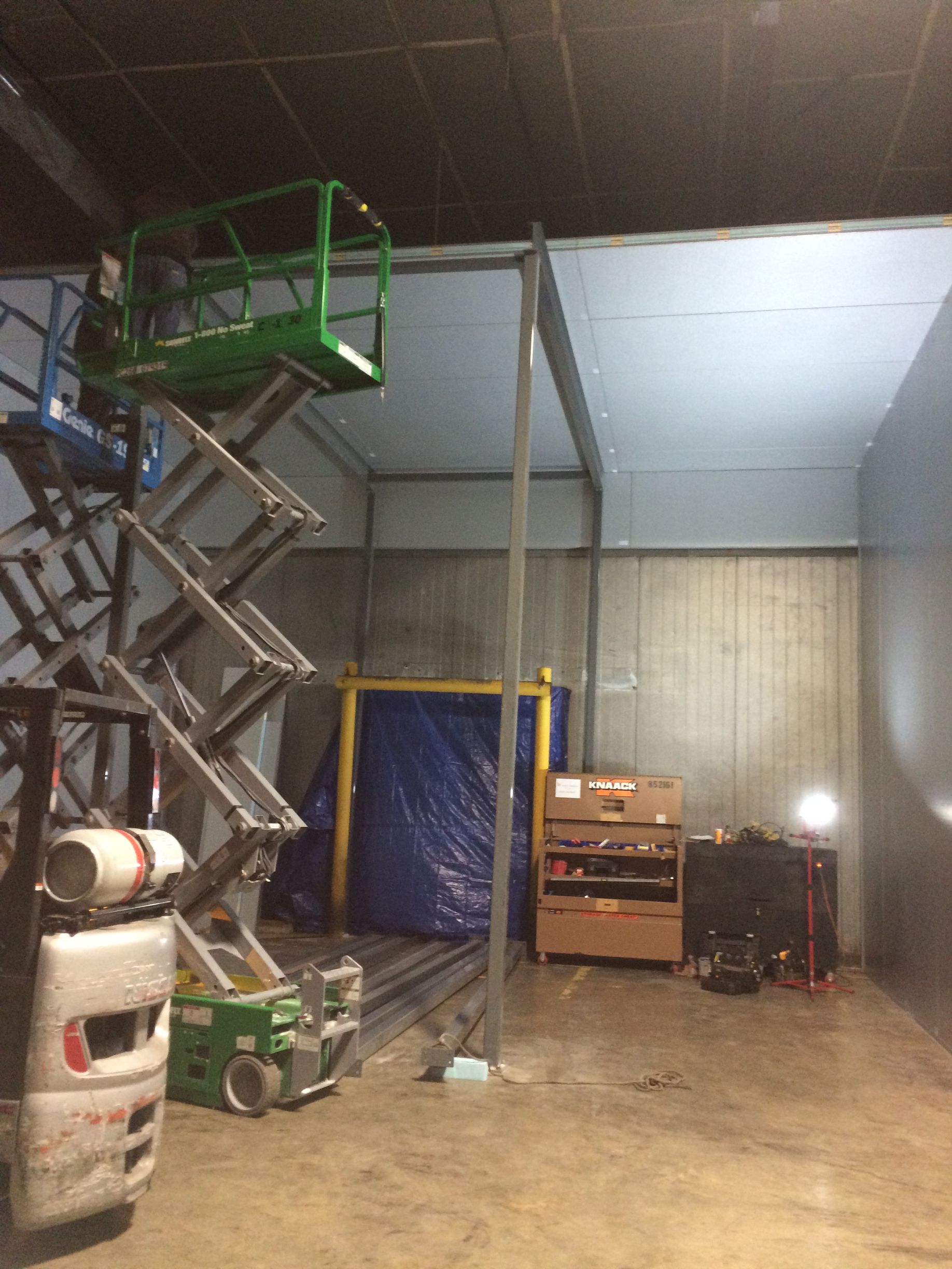 Industrial warehouse interior with a scissor lift, ladder, blue tarp, and equipment under high ceiling lights